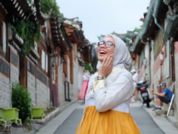 A young woman wears a gold skirt that flows from a white bodice and long sleeves. Her left hand is touching her face. She looks delighted.